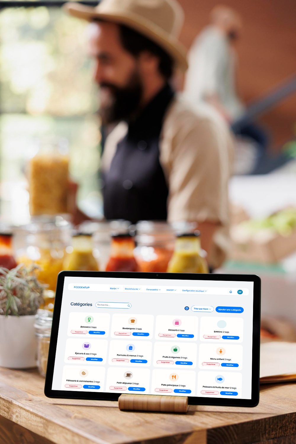 Tablette posée sur un comptoir affichant la gestion des catégories de la carte FoodEatUp, commerçant en arrière-plan.